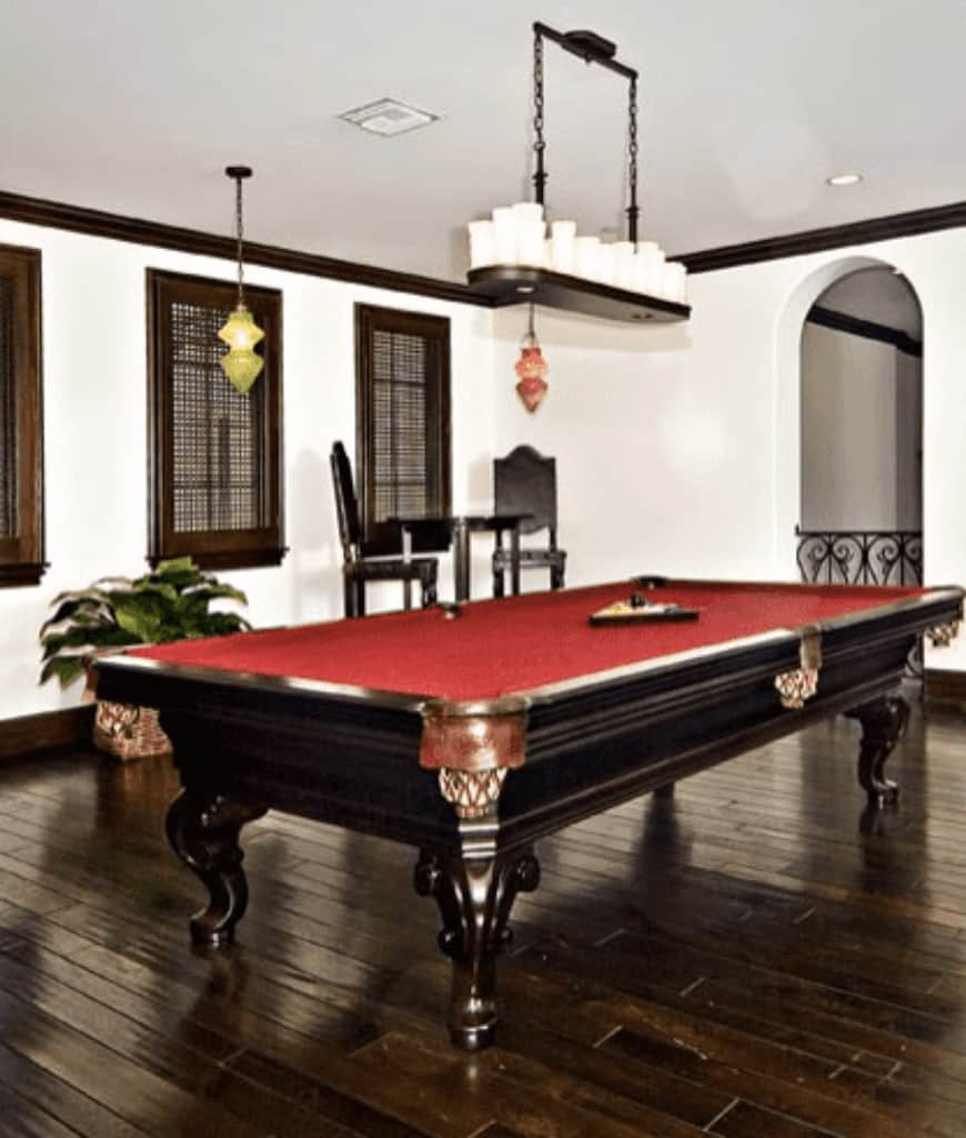 经典的白色房间设有一个红色台球桌，由线性吊灯照明，并伴随着木制框架窗的黑色酒吧椅。