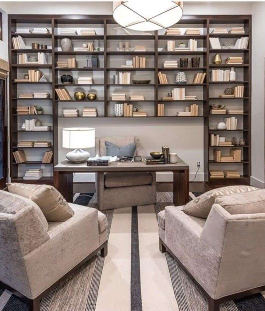 在这间家庭办公室里，一对灰色扶手椅正对着一张放在条纹地毯上的木桌，开放式书架上摆满了书籍和花瓶。