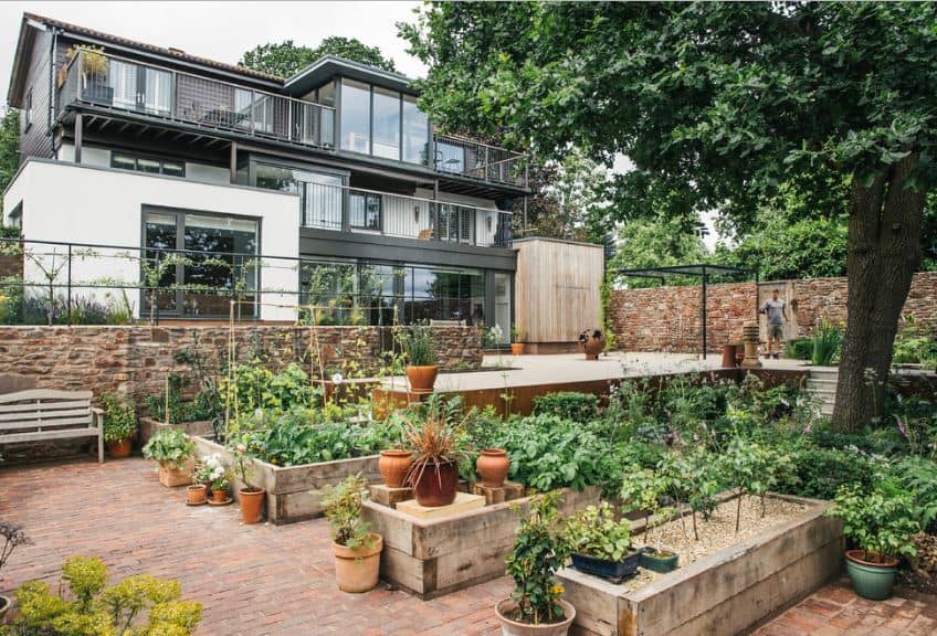 这座现代的房子充满了玻璃墙和阳台，在房子的前面有一个广阔的花园。在这个花园的前面是一个开阔的户外聚会区，通往花园的盆栽植物和砖地板的小楼梯。