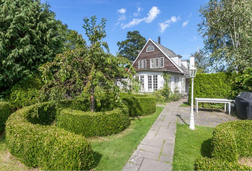 这是一个斯堪的纳维亚风格的景观前院的木制简单的房子。这里的景观设计得很好，看起来像一个公园，有灰色的石头人行道，树木被精心修剪的灌木树篱包围，甚至还有一根公园灯柱。