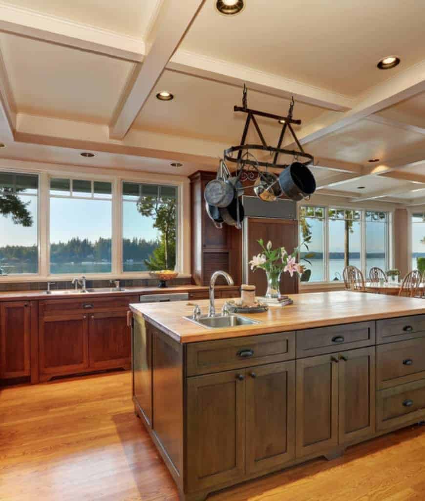 温暖的厨房，木橱柜和全景窗户俯瞰风景优美的室外景色。它包括一个深色的木质早餐岛架和挂在格子天花板上的老式锅架。