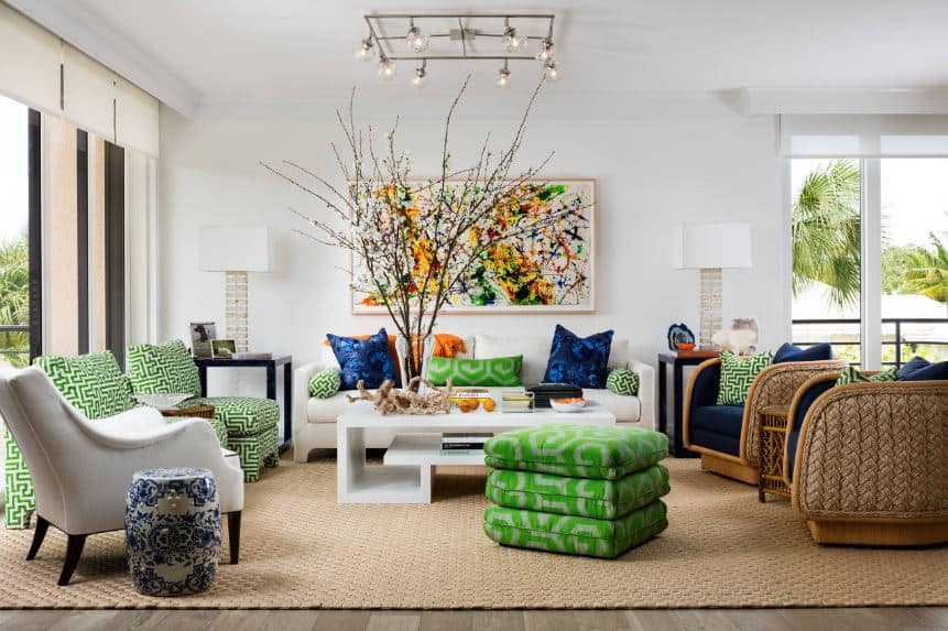 热带风格客厅功能丰富多彩的溅靠垫、绿色和蓝色的枕头,和椅子。家具,包括一个沙发软垫和编织柳条椅子,白色与绿色和蓝色的口音的颜色对比。设置在一个编织地毯、家具面临着高雅几何咖啡桌,虽然同样丰富多彩的抽象画挂在墙上在沙发后面。大窗户照亮明亮的空间,增强当代吊坠灯具,台灯。