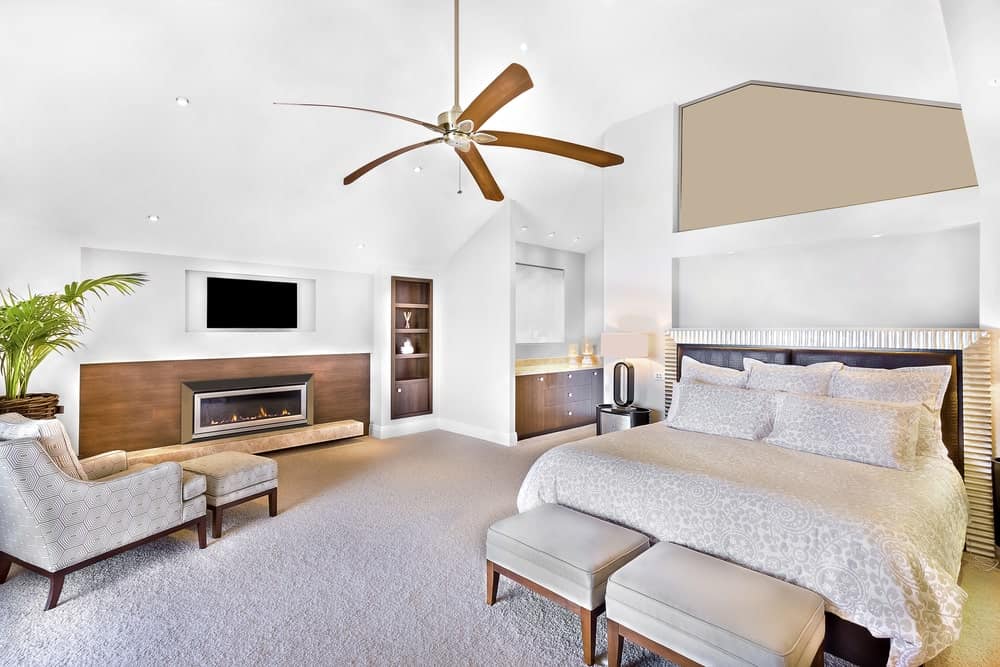 这是一间白色的主卧室，强调了木材和灰色色调的元素。白色的墙壁和天花板与浅灰色的地毯地板和优雅的壁炉相搭配，壁炉周围的木板设计与旁边的内置架子相匹配。