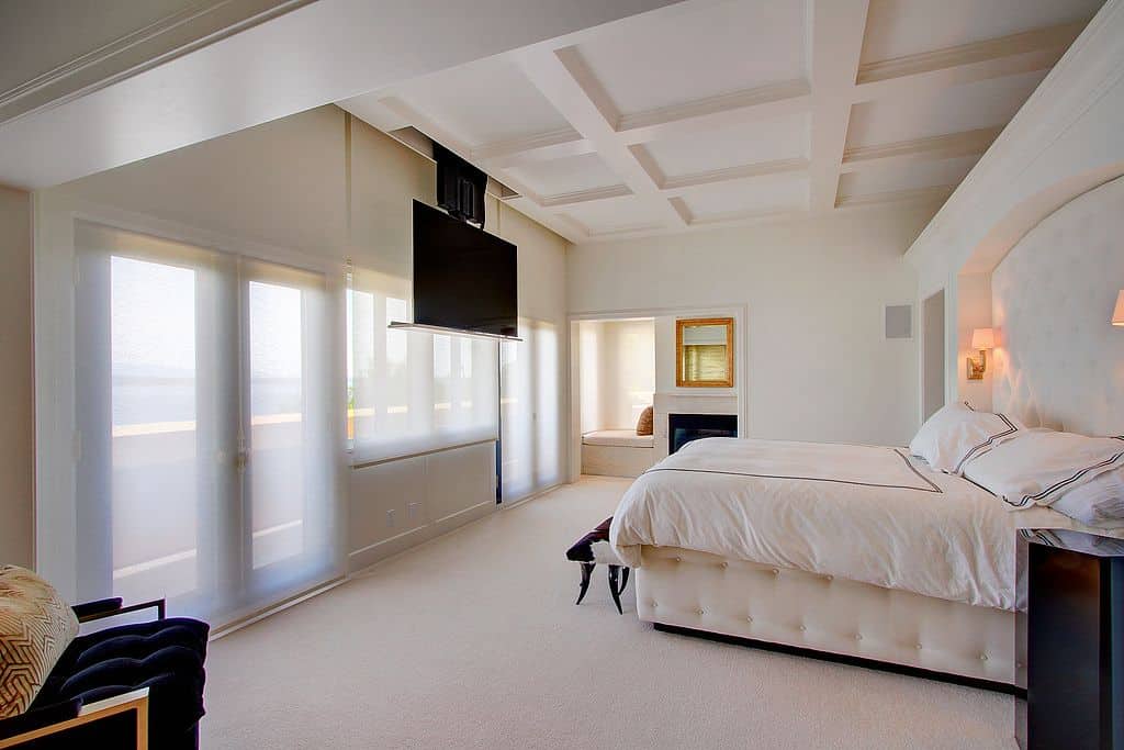 时尚的主卧室，白色墙壁和白色地毯地板。床上有两盏带暖灯的壁灯。卧室侧面有一座壁炉，床前的天花板上挂着一台宽屏大电视。