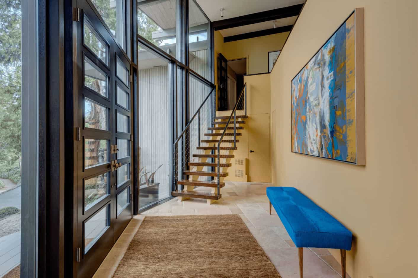 当代的门厅强调了一个明亮的蓝色长凳放置在巨大的墙壁艺术。它有一个现代化的楼梯和通高的玻璃俯瞰宁静的户外景观。