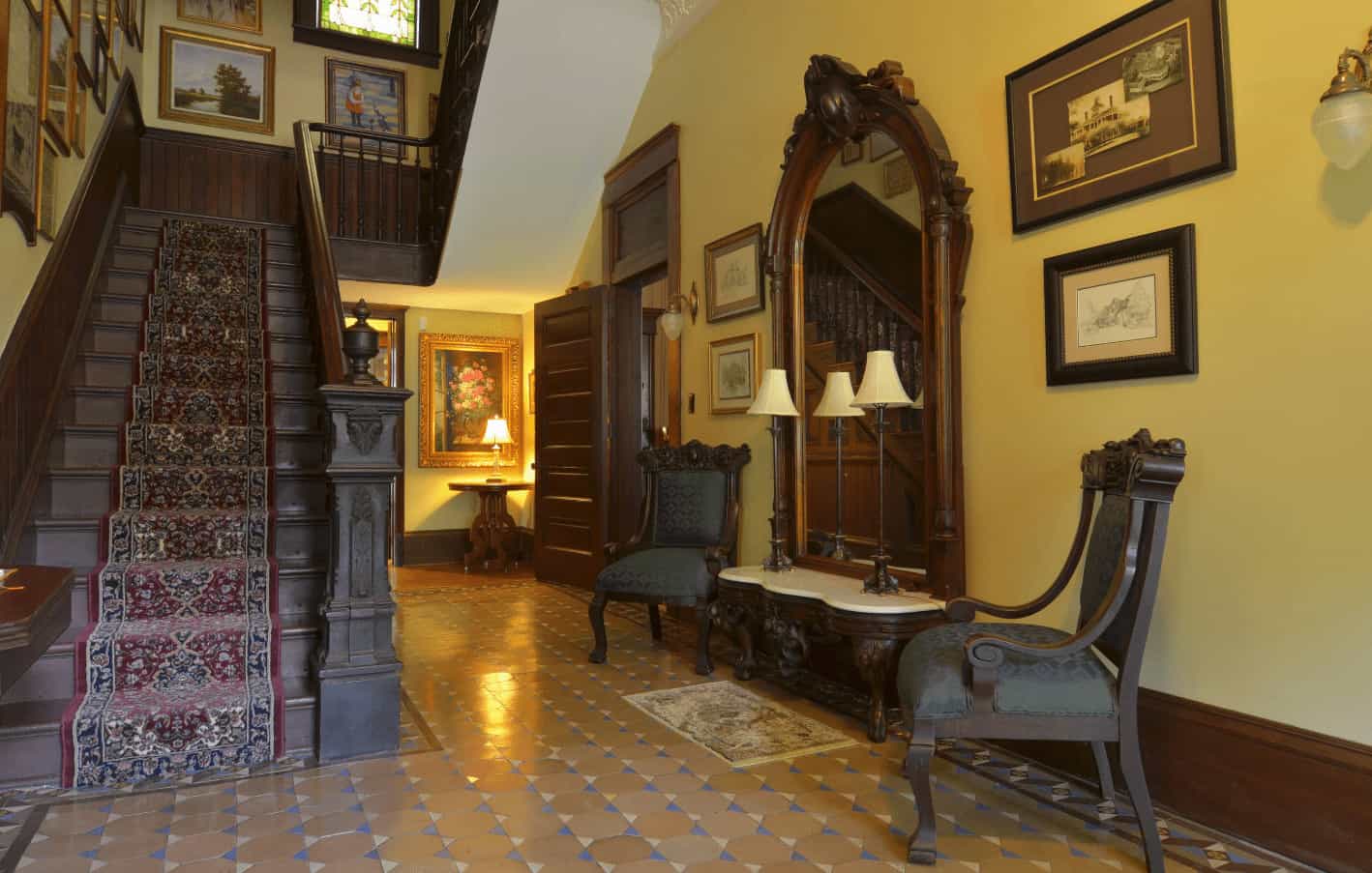 维多利亚式门厅拥有一个雕刻的木头控制台桌子，配以一面巨大的镜子和深色的木头扶手椅。它有黄色的墙壁，上面挂满了裱框的墙壁艺术作品，还有一个木制楼梯，上面放着一个老式的跑步机。