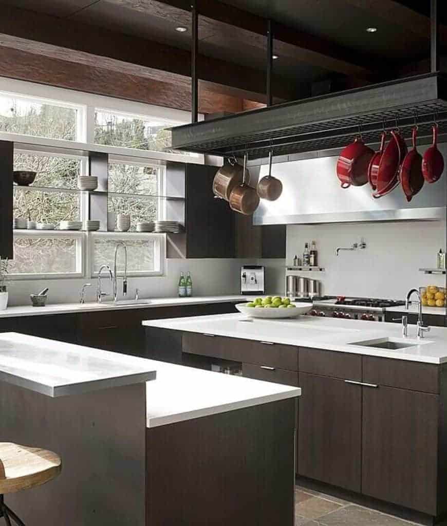 现代厨房拥有一个巨大的金属锅架，挂着棕色和红色的锅，悬挂在两层早餐柜台对面的深色木厨房岛上。