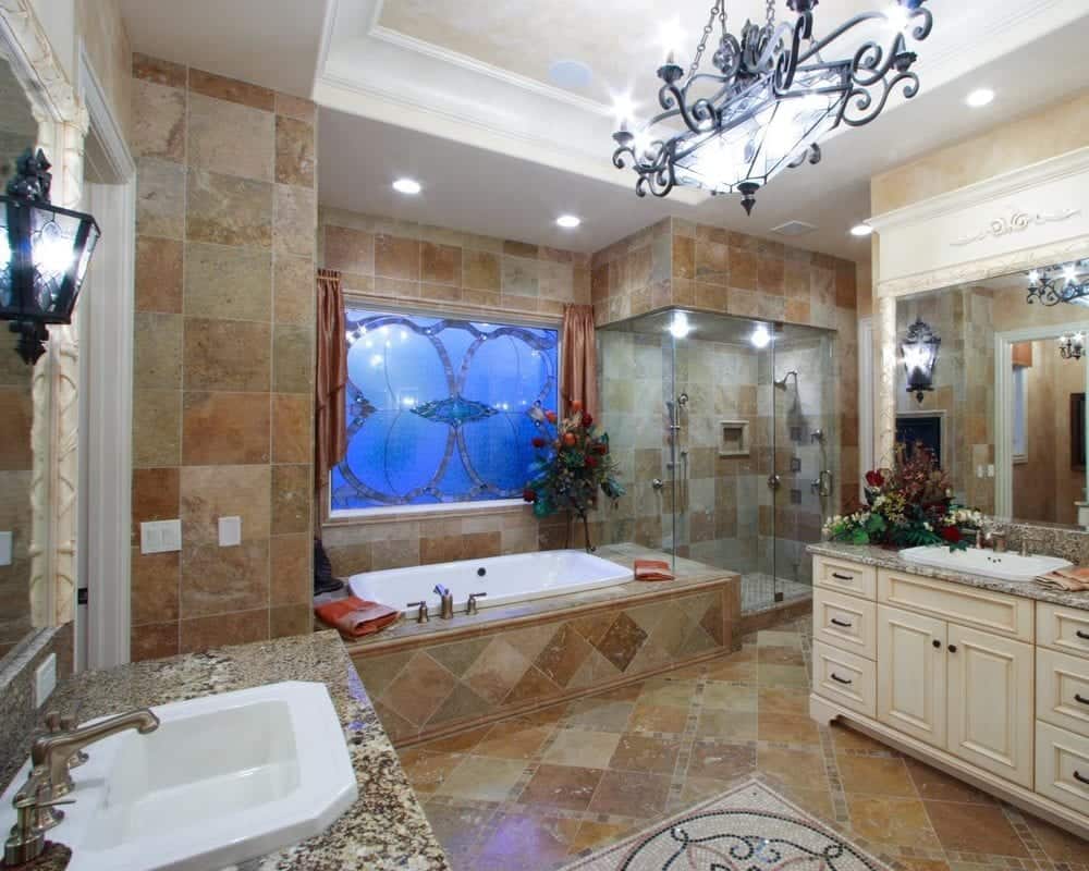 这是一间主浴室，地面的泥土瓷砖上方挂着一盏锻铁枝形吊灯。这些瓷砖也被用来安置在玻璃淋浴区旁边角落的浴缸。