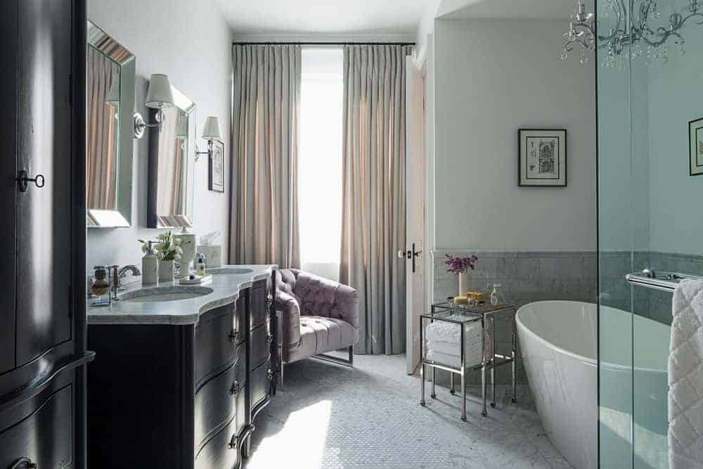 过渡风格的浴室，有水晶吊灯，独立浴缸，在瓷砖地板上的双洗手池旁边有一个座位。