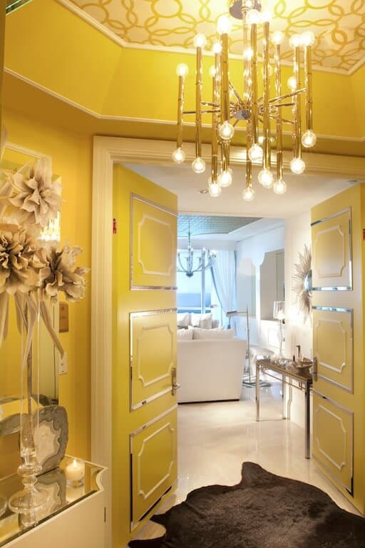这个优雅的门厅以明亮的黄色墙壁和凹形天花板为特色，明亮的金色吊灯在白色地板上投射出温暖的黄色灯光，地板上覆盖着棕色的动物皮毛区域地毯。