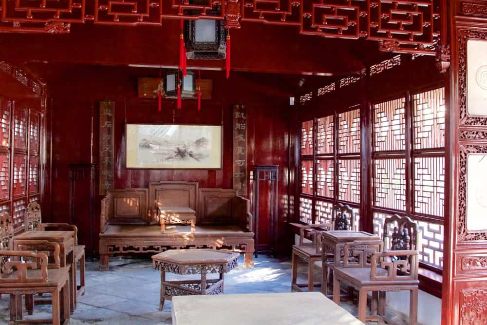 这是一间传统的中式客厅，窗户上有东方图案，与中间小桌子周围的扶手椅和木凳上的雕刻相匹配。红色的木制天花板上挂着中国灯笼。