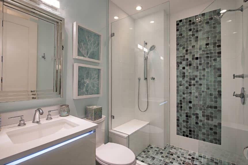 这个简单的海滩风格浴室的墙壁和玻璃围成的淋浴区有各种深浅的灰色，淋浴区旁边是白色马桶，上面有绿色艺术品，旁边是一个带LED照明的现代灰色梳妆台。