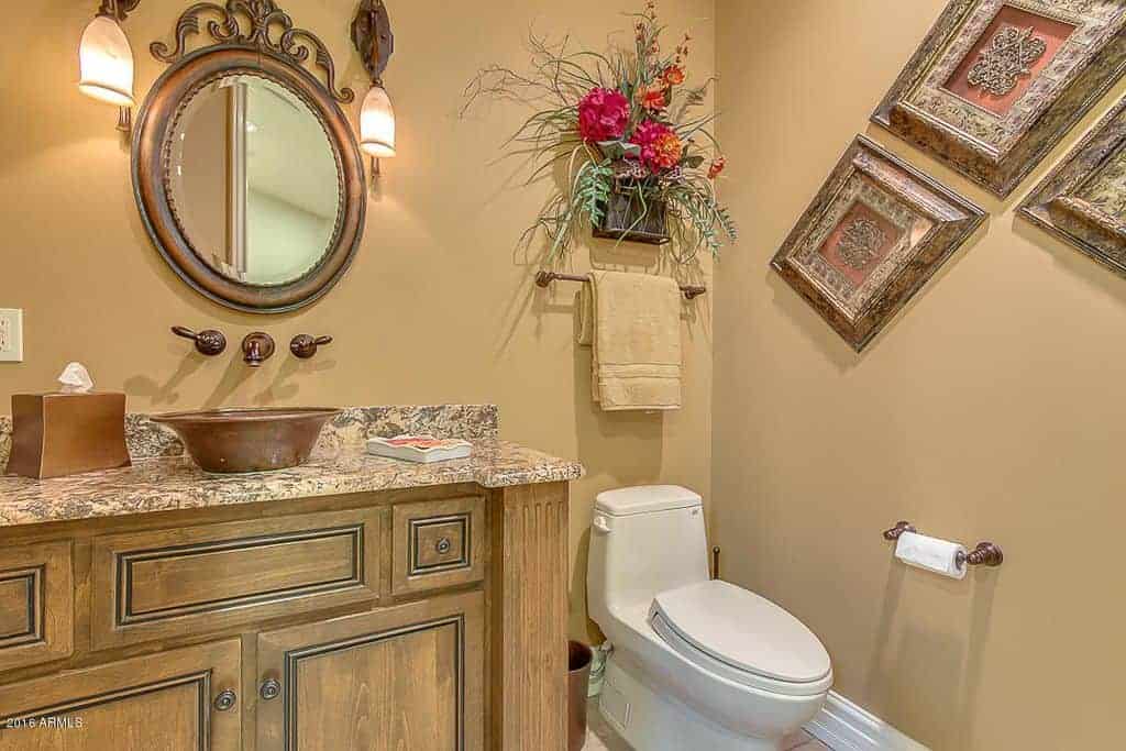 华丽的浴室设计有花卉和华丽的墙壁艺术，还有一面圆形镜子，挂在木制梳妆台上方，上面有大理石柜台和青铜器皿水槽。