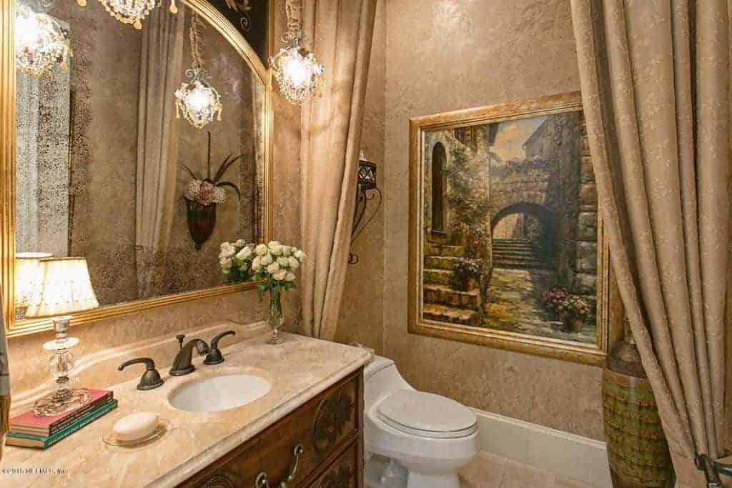 水晶吊灯照亮了这个米黄色的浴室，展示了一个大理石顶部洗手台和一个可爱的画框强调的厕所区域。