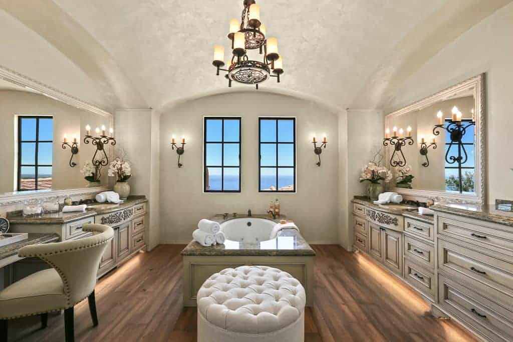 这间主浴室的特色是面向水槽的梳妆台，中间有一个浴缸，旁边有一个白色的簇绒软凳，坐落在天然木板地板上。