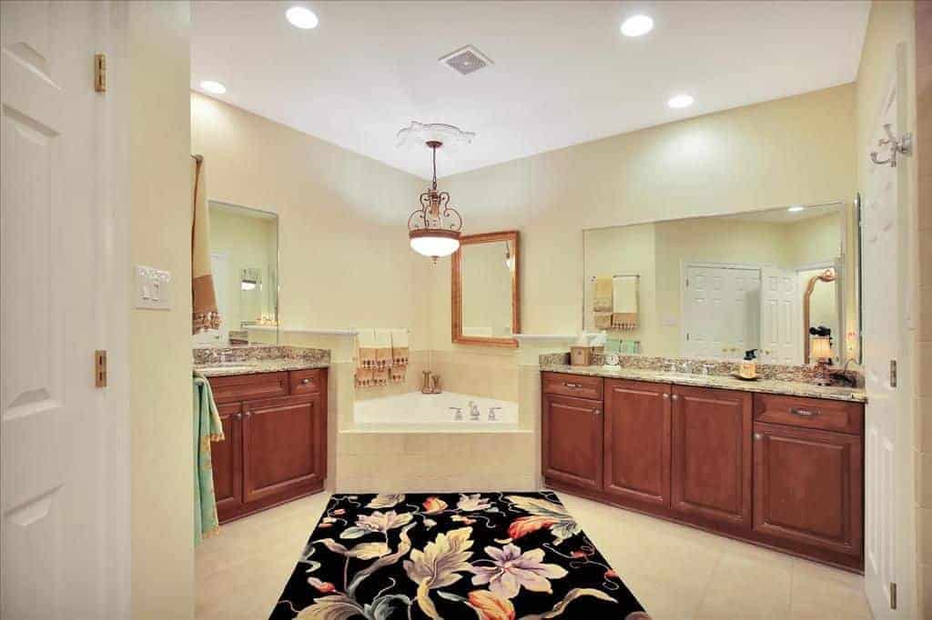 华丽的黑色碎花地毯在这个米色的主浴室中脱颖而出，展示了木质梳妆台之间的角落浴缸，由一盏华丽的吊灯照亮。