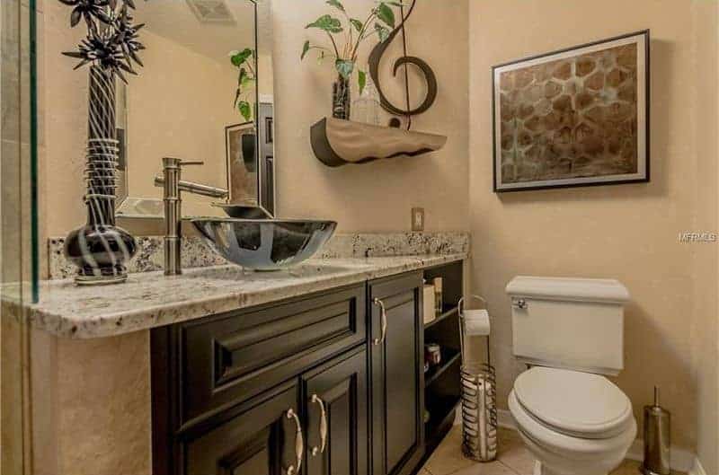 在这个米色的浴室里，一幅有趣的墙上艺术品悬挂在厕所上方，有一个时尚的架子和一个深色的木质梳妆台，上面有一个镀铬的水槽和华丽的黑色花瓶。