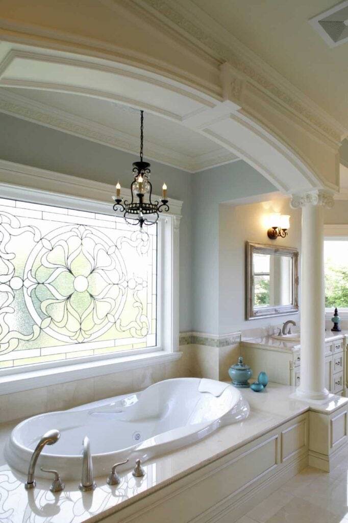 优雅的浴室，洗手池梳妆台和凹壁浴缸下的迷人的彩色玻璃窗内衬白色柱。它由一盏锻铁枝形吊灯和安装在华丽镜子上方的壁灯照明。