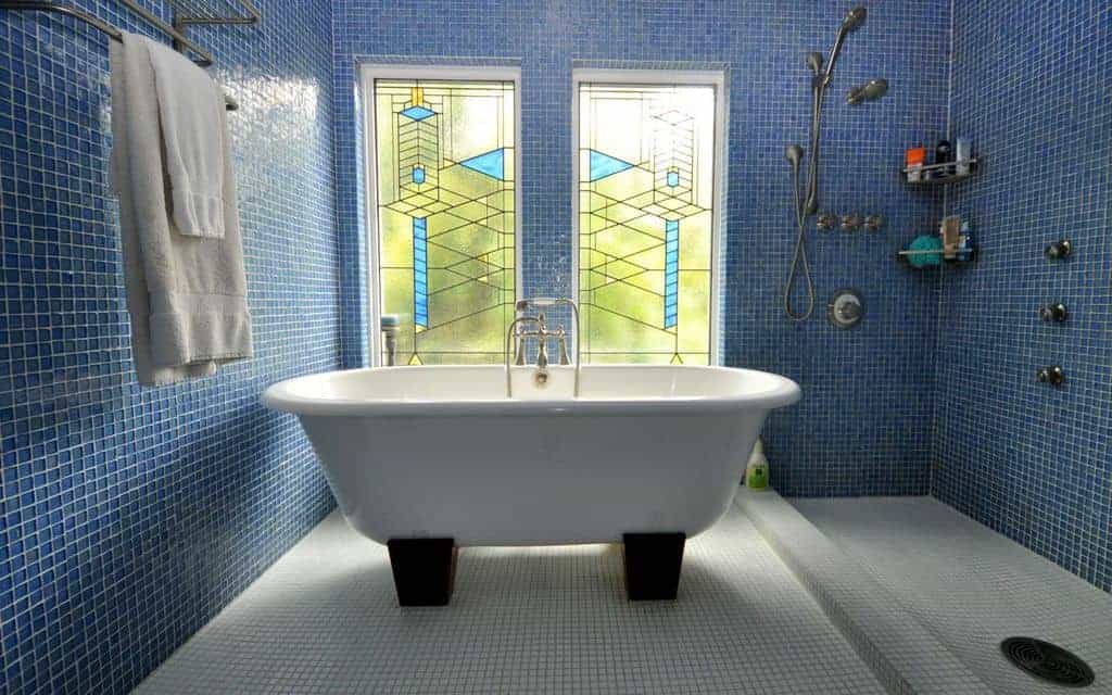 迷人的浴室，开放式淋浴区和基座浴缸，周围环绕着神话般的彩色玻璃窗和蓝色瓷砖墙壁，配有铬固定装置和角落架子。