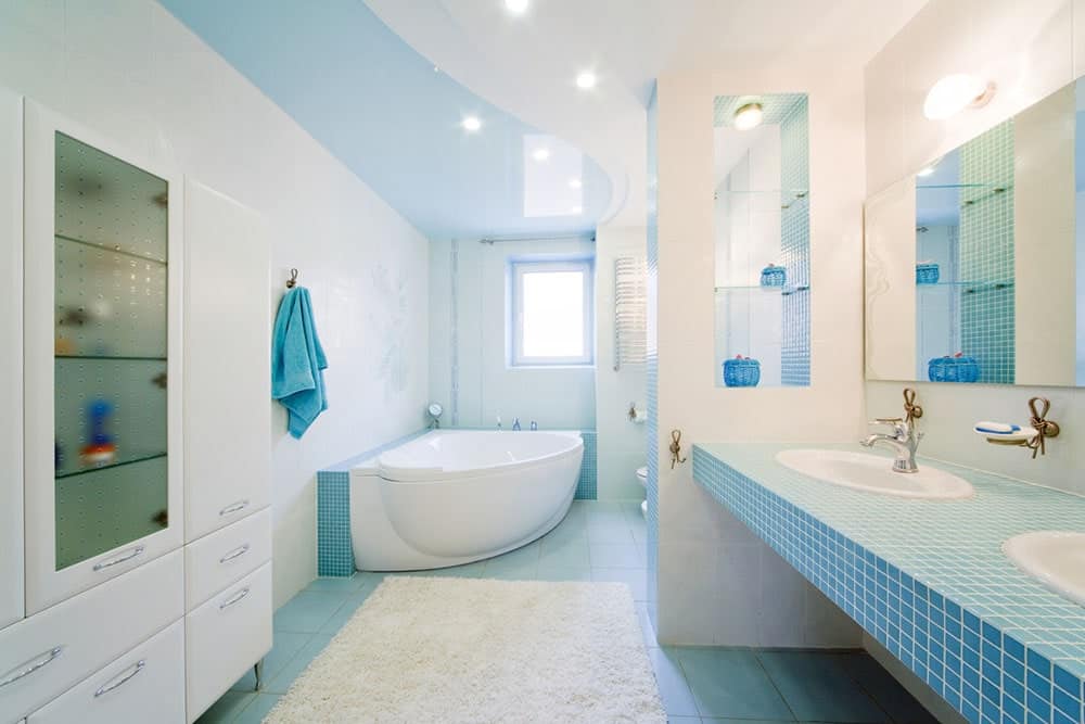 明亮的浴室拥有一个落地式浴缸和光滑的白色橱柜，横跨蓝色瓷砖台面，配有双水槽和铬水龙头。