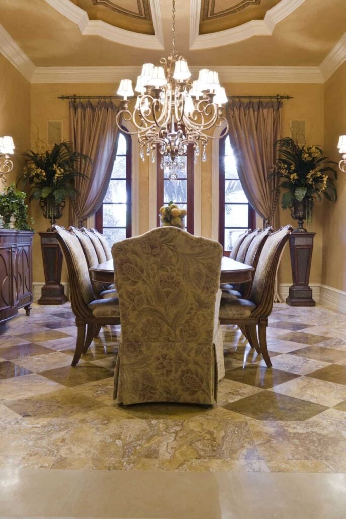 这间餐厅铺着深浅不一的棕色大理石地板，餐椅上有棕色图案的滑套，米黄色墙壁上有窗帘和装饰品。