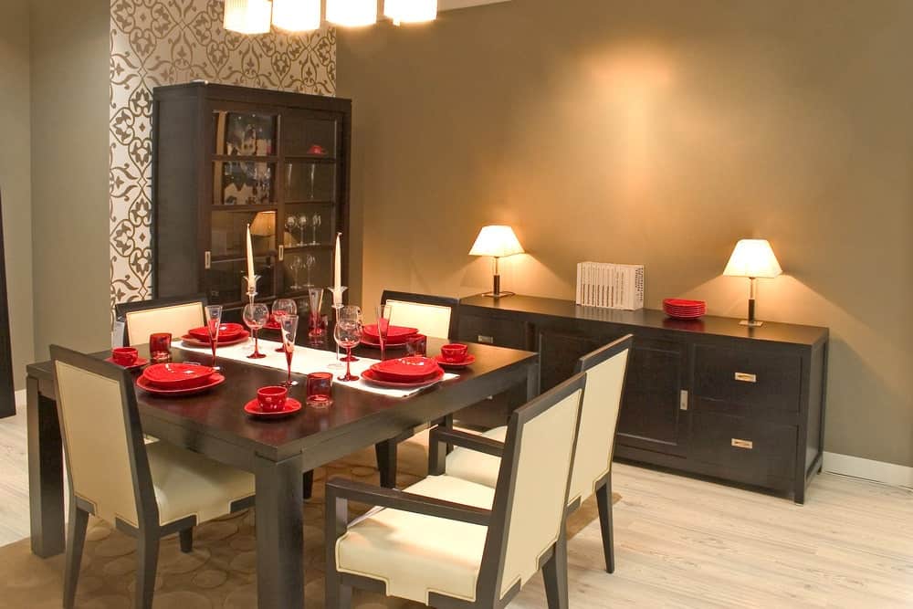 红色的陶瓷餐具与深棕色的木桌形成了鲜明的对比，与米色皮革靠垫的木制餐椅完美搭配。这也与低矮的餐厅橱柜相匹配，在灰色的墙壁上有两盏台灯。