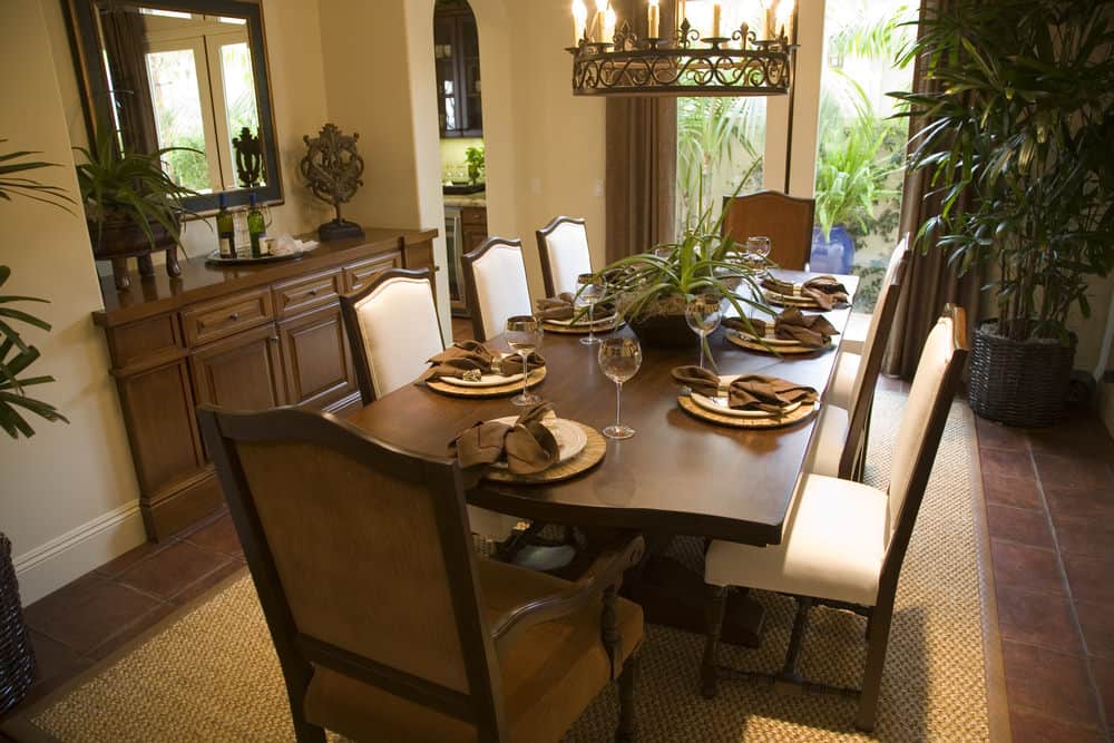 农场风格的圆形吊灯有着深棕色的华丽设计，与优雅的深棕色桌子的美学相匹配，餐厅的橱柜嵌入米黄色的墙壁中，与之形成对比的是与陶土地板相匹配的深棕色窗帘。