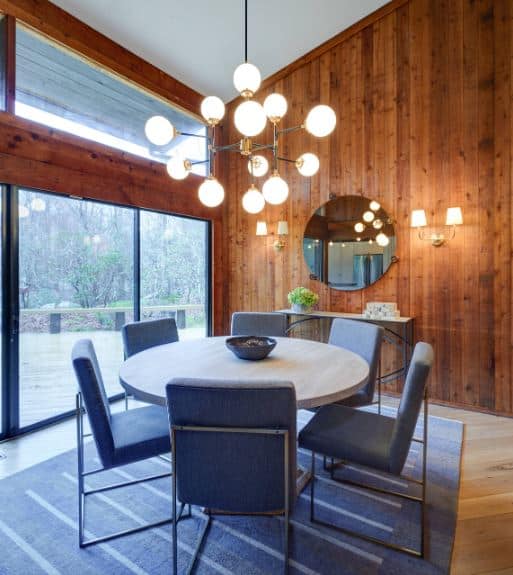白色的棚顶与木墙形成对比，木墙在控制台桌子上方装饰着壁挂式圆镜子，两侧的壁灯与浅灰色餐桌上方的吊灯相匹配，餐桌上配有蓝色软垫现代椅子。