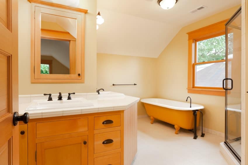 米黄色角落里的独立浴缸的橙色色调与木质l形洗手台的橙色色调相匹配，洗手台与角落的布局相匹配，洗手台的工作台面在淋浴区玻璃门对面铺有白色瓷砖。