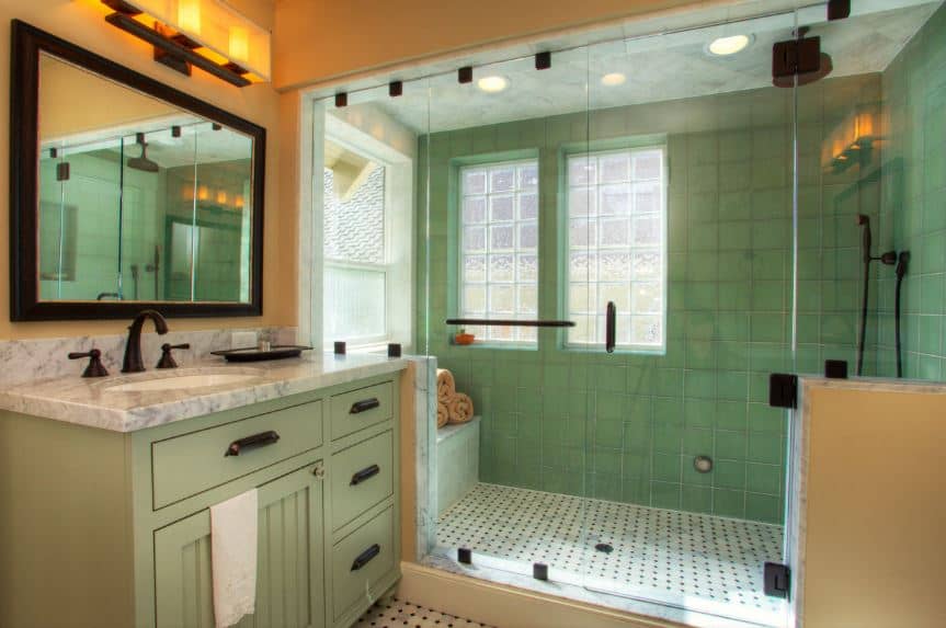 远处墙上的大型玻璃围淋浴区墙上贴着绿色瓷砖，与梳妆台上的牛油果绿色橱柜和抽屉完美搭配，梳妆台的黑色把手与浴室的固定装置相匹配。