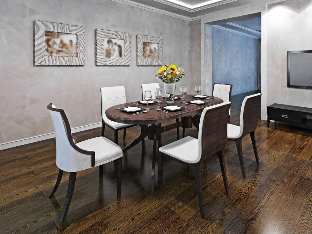 白色软垫椅子坐在椭圆形餐桌旁，这个简单的用餐区有硬木地板和混凝土墙，墙上挂着可爱的相框照片。