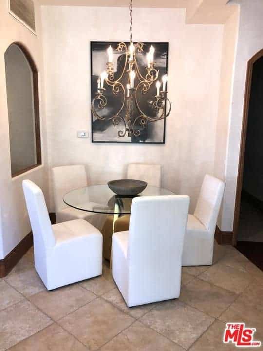 一个小的用餐区，有光滑的白色椅子和一个玻璃餐桌，上面有一个装饰性的碗。它包括一盏华丽的枝形吊灯和一幅安装在白墙上的抽象艺术品。