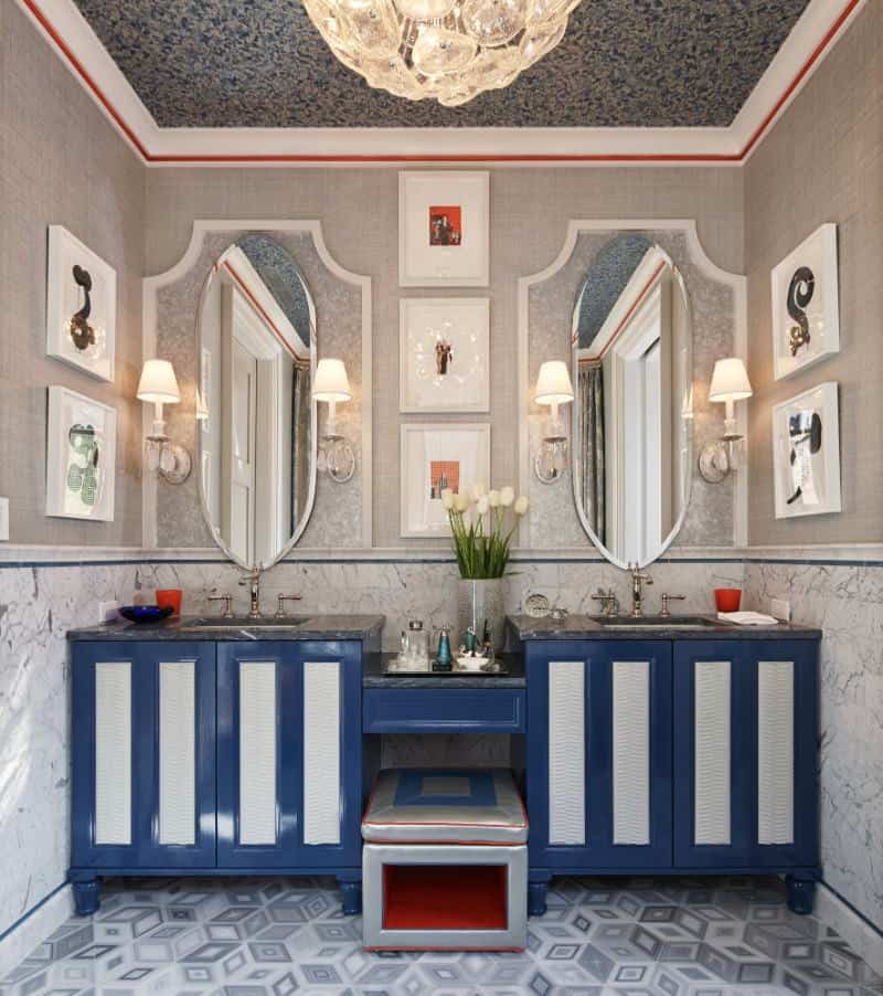 兼收并蓄的化妆室装饰着白色框架的墙壁艺术和椭圆形的镜子，镜子安装在蓝色的洗手台上方，旁边还有一个灰色的软垫凳子。