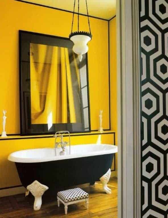 黄色的浴室里有一面大镜子，两侧是迷你雕塑，还有一个圆点的凳子和一个白色玻璃吊坠照亮的黑色爪足浴缸。