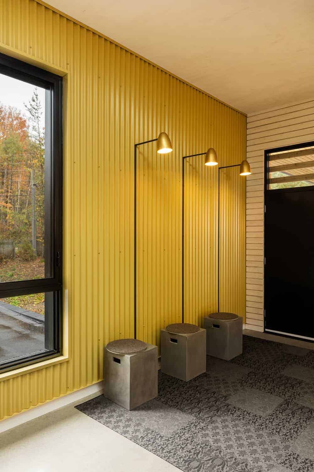 这个简单的工业风格的门厅在白色地板上有灰色图案的地毯。令人愉快的黄色金属墙壁和壁挂式灯具使这个空间更加明亮，这些灯具安装在三个内置的混凝土小凳子上。