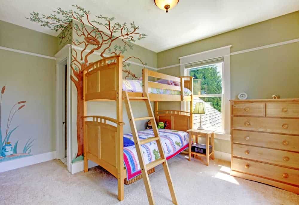 孩子们的卧室里有一张木制双层床，抽屉柜和一个床头柜，上面有一盏传统的台灯。它有地毯地板和灰褐色的墙壁，墙壁上有植物和树木的贴纸。