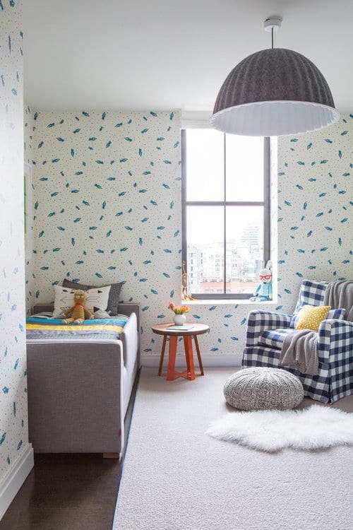 这间卧室铺着蓝色的鱼壁纸，展示了一张灰色的床和格子裙边的椅子，由一个巨大的圆顶吊坠照亮。