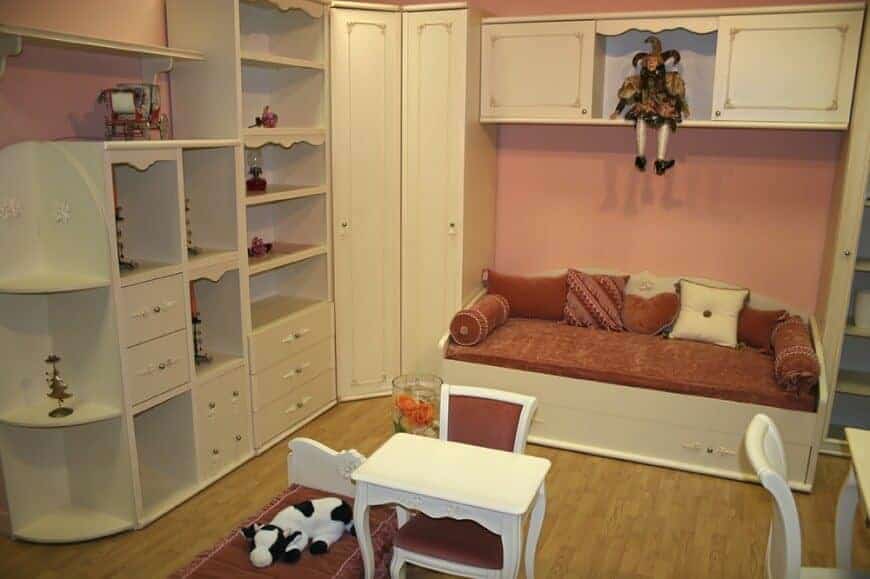 在这间儿童卧室里，一个小丑玩偶坐在浮动的柜子上，里面有内置的储物柜和架子，还有一张白色的床，上面有红色的床垫和枕头。