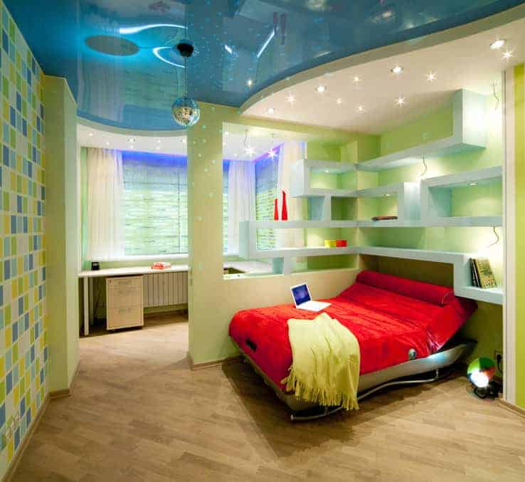 华丽的卧室，硬木地板和时尚的天花板安装有嵌入式灯和铬球吊坠。房间里有一张红色的床和一张l形的桌子，中间用内置的书架隔开。