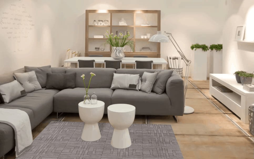 这个起居空间以棕色地板和白色墙壁为特色。它有一个l型的灰色沙发和一个灰色的时尚地毯，上面有两张白色的中央桌子。