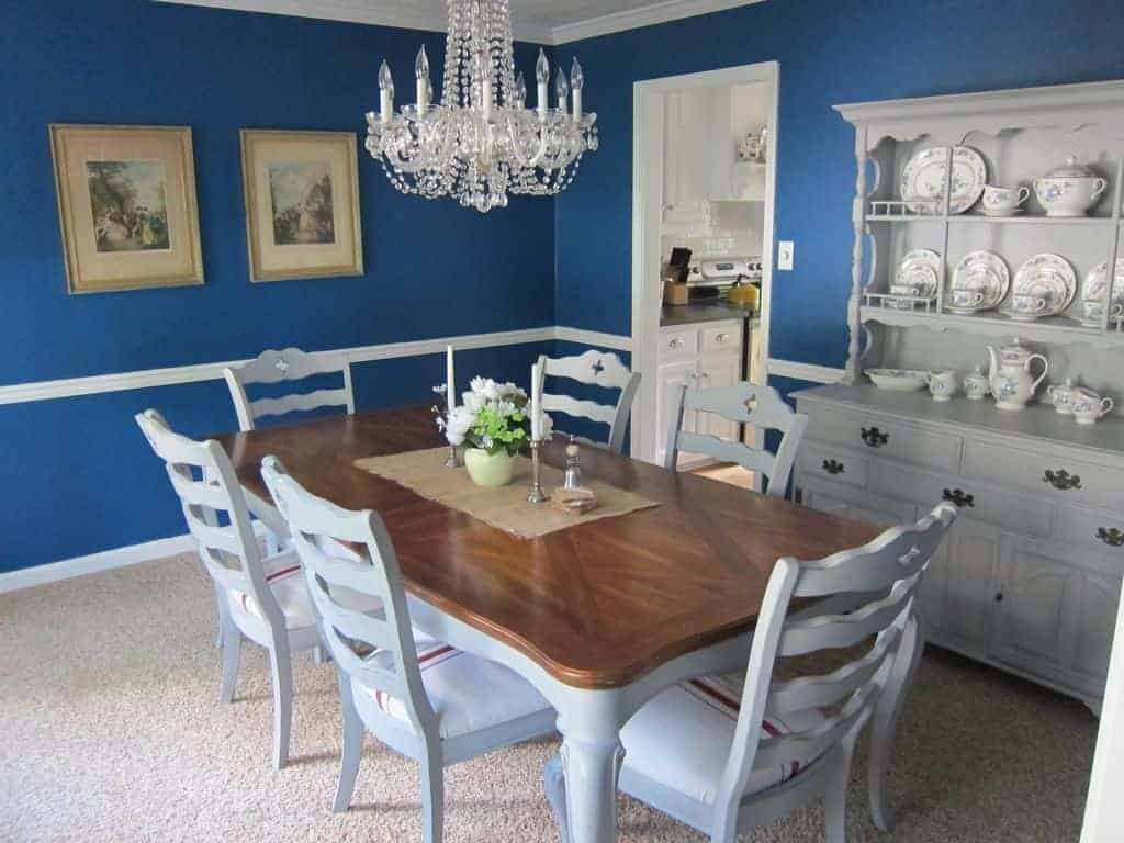 木餐桌周围的白色木制椅子有一波设计其回来,与亮蓝色的墙上装饰着两个经典的绘画和浅灰色餐厅内阁与中国一套别致的餐具。