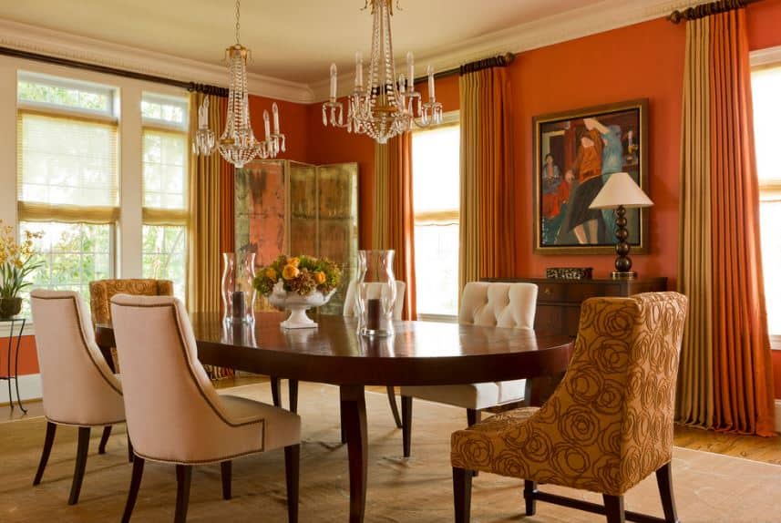 大木制餐桌的头部有玫瑰图案的米色椅子，其余的则是白色软垫。这与高橙色的墙壁搭配，高窗户有橙色和米黄色的窗帘，以完成美学。