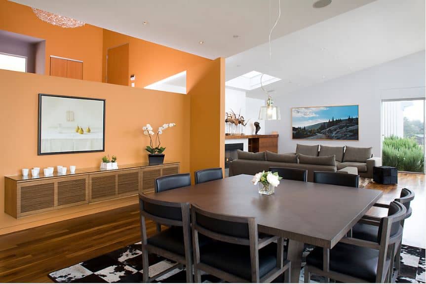 方形餐桌的深灰色色调与周围的椅子相匹配，黑色皮革靠垫与下面的花纹地毯相匹配。橙色的墙壁及其装饰的欢快风格软化了这一点。