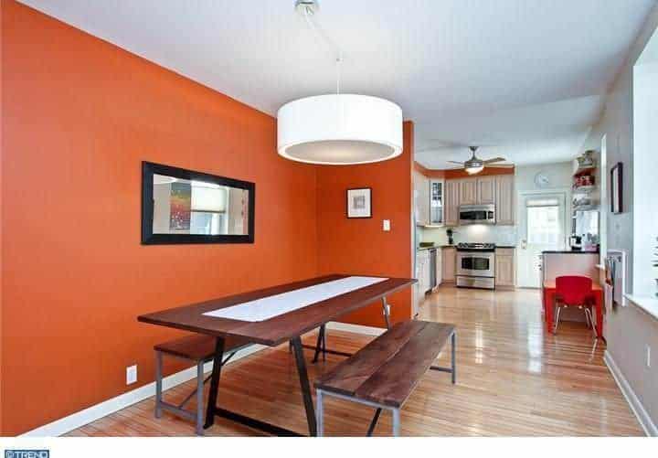 这个简单的餐厅有一个极简主义的氛围，它的简单的木制矩形桌子和两个侧翼的木制长凳上有一个圆形的白色吊灯。这是一个温暖和舒适的背景与泥土的橙色阴影。