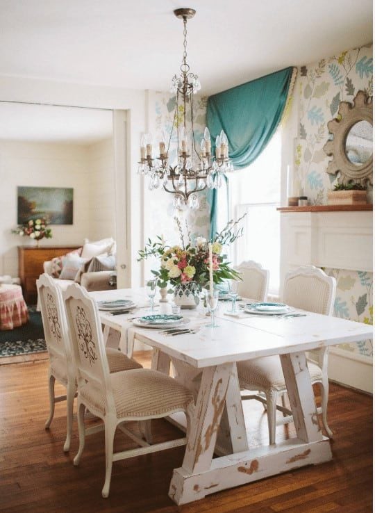 华丽的餐厅，软垫椅子和一个破旧的白色餐桌由蜡烛吊灯照亮。它铺着木地板，窗户上挂着可爱的蓝色窗帘。