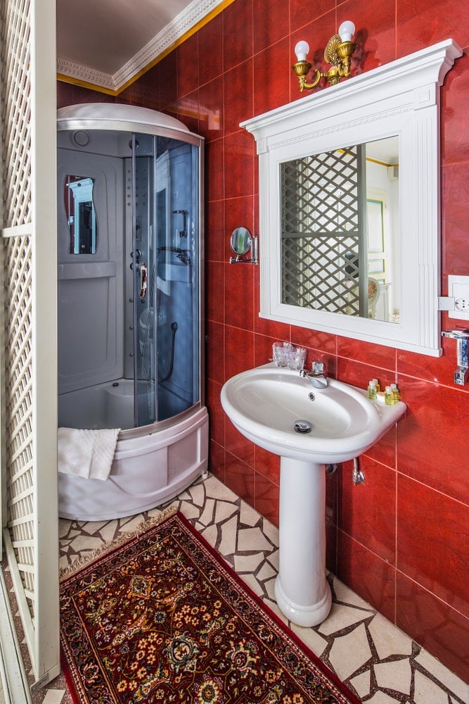 这间小而简单的西南风格浴室墙壁上有土红色的瓷砖，地板上有白色马赛克瓷砖的红色图案地毯。这面铁锈色的墙壁使白色的瓷底座水槽以及旁边的淋浴区脱颖而出。