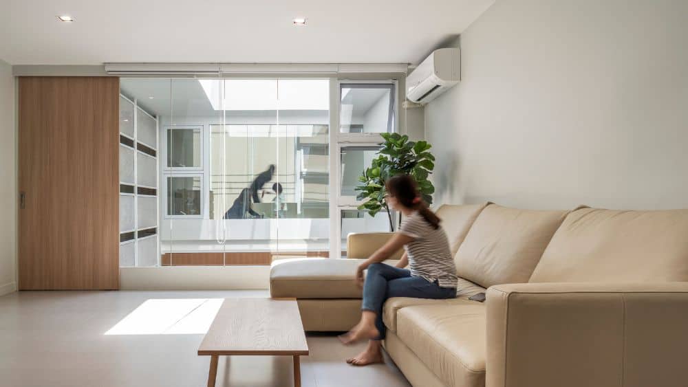 这是一个简单的客厅，米黄色的大l型沙发搭配木制咖啡桌，大玻璃墙旁边的角落里摆放着盆栽植物。
