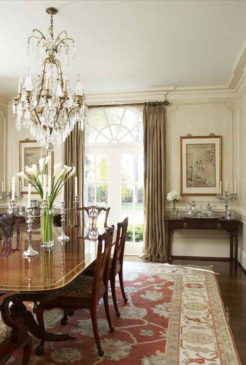 白色的餐厅有通高的护墙板和一扇覆盖着灰褐色窗帘的法式门。它包括一盏漂亮的枝形吊灯和一套放在红色地毯上的高级餐具。