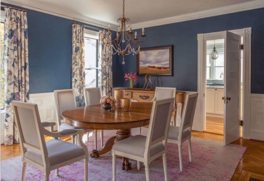 维多利亚时代的餐厅展示了一个铜吊灯和景观墙艺术安装在蓝色的墙壁上，覆盖着白色的护墙板。它包括一张木制餐桌和白色软垫椅子，上面铺着一张破旧的粉色地毯。