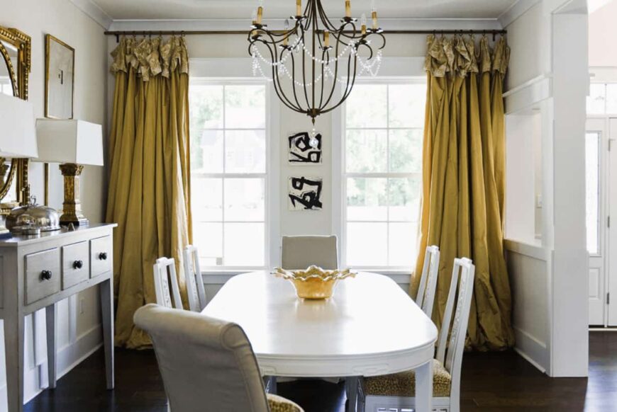 白色的用餐区以金色的镜子和黄色的窗帘覆盖着白色的框窗。里面有一盏复古的枝形吊灯，有软垫的椅子围绕着一张椭圆形的桌子，桌子上有一个装饰性的碗。